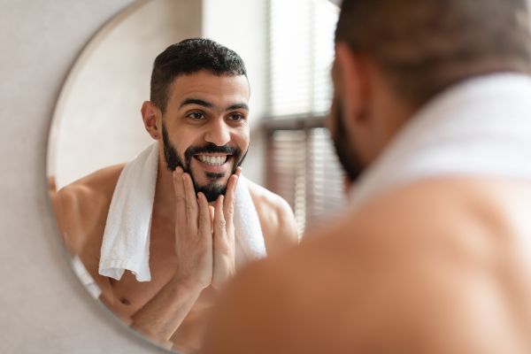 Minoxidil barba; presentación de un plano medio corto de un hombre mirándose al espejo mientras visualiza los resultados en su barba tras utilizar un tratamiento.
